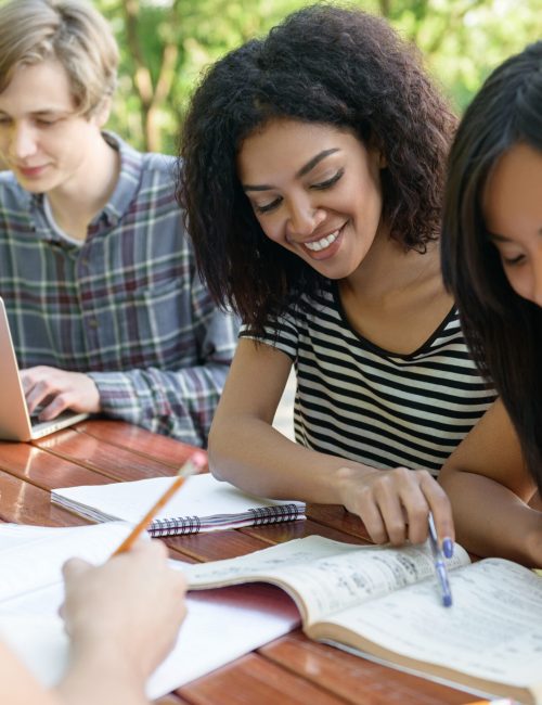 Image of multiethnic group of young happy students sitting and studying outdoors while talking. Looking aside.